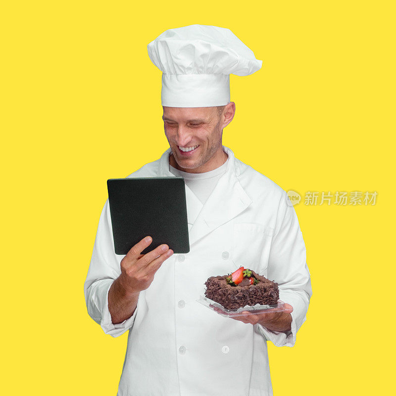 黄色背景前是白种男厨师，穿着夹克衫，手持蛋糕，使用数码平板电脑