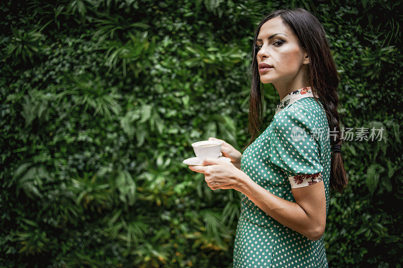 一个美丽的女人站在一堵绿色的墙和喝咖啡-一个优雅的女人旁边的天然树叶墙