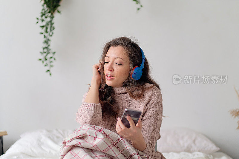 年轻快活的女人躺在床上，用手机拖着脚唱着歌。放松的女孩在家里用耳机听音乐。快乐的女人在不经意间享受着卧室里的音乐，同时发着手机短信