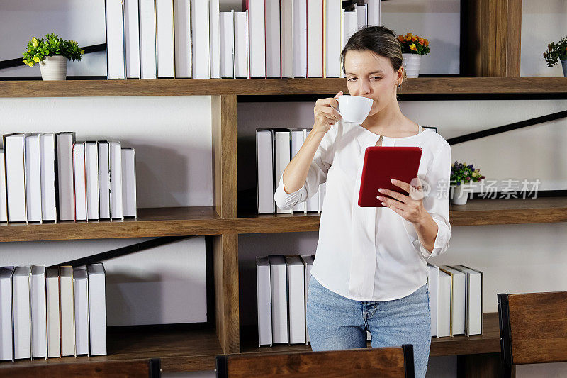一名微笑的白人女子正在图书馆喝咖啡，在平板电脑上阅读在线新闻以获取知识，并在大学或现代办公室利用互联网工作。科技教育理念