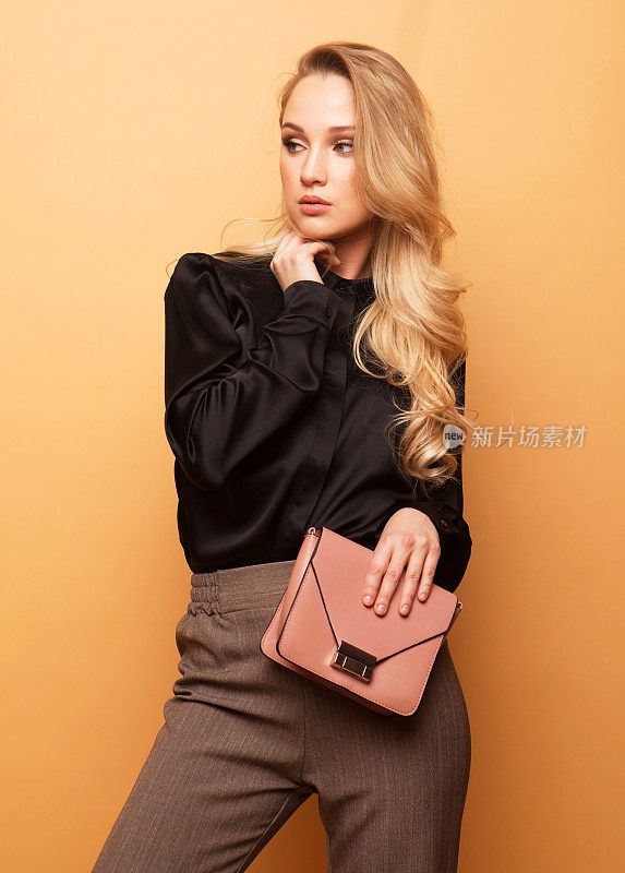 年轻漂亮的金发女人穿着黑色的衬衫和棕色的裤子，拿着一个粉红色的手提包