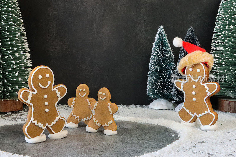 三个自制的姜饼人在池塘上滑冰的特写图像，姜饼人戴着圣诞帽，下雪了，夜晚的圣诞森林场景与模型树，驯鹿和圣诞树饼干，黑色的背景