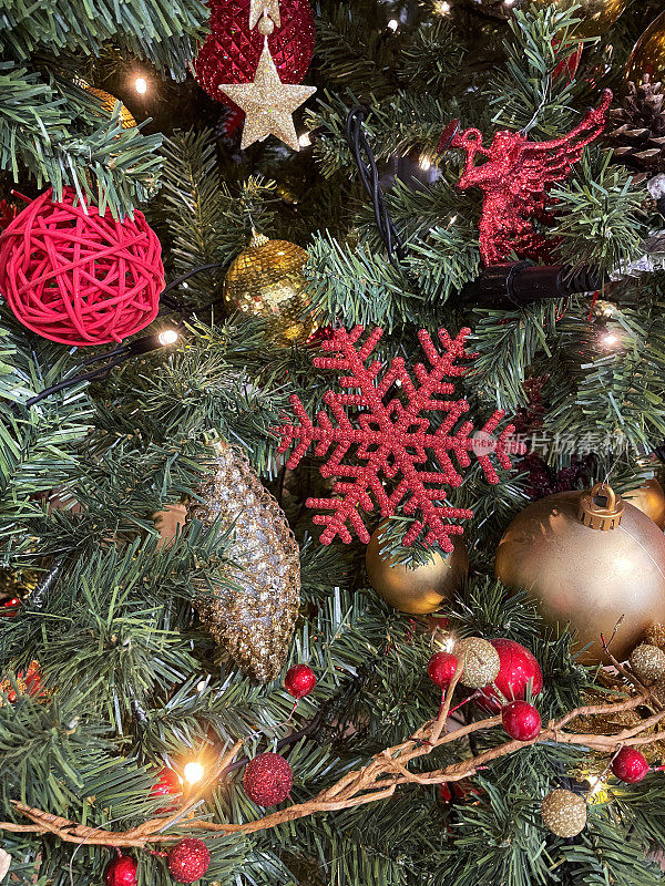近距离拍摄的红色闪光雪花挂在圣诞树的云杉针叶上，周围环绕着红色和金色的小饰物，模糊的白色仙女灯背景，前景聚焦