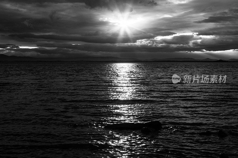 意大利翁布里亚特拉西梅诺湖上的日落，太阳落在忧郁的云层后面，美丽的水面倒影