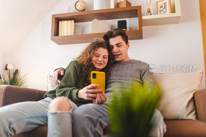 美丽的年轻夫妇在客厅放松，同时看媒体在他们的智能手机