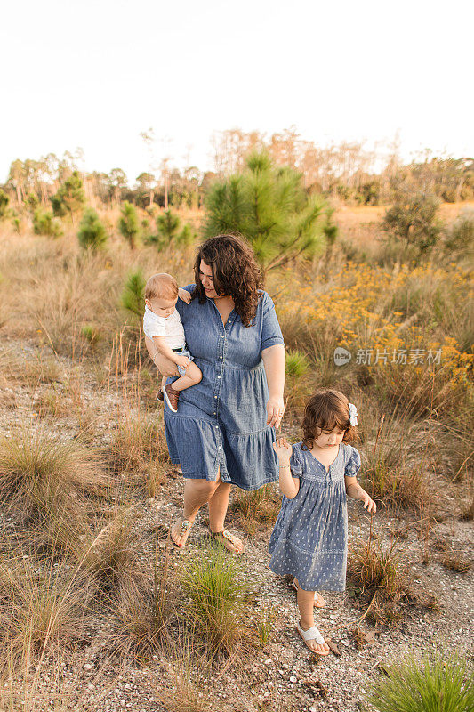 日落时，一位31岁的母亲和她三岁的古巴裔美国人女儿，棕色卷发和棕色眼睛，穿着蓝色连衣裙，抱着她的男婴，穿着白色Polo衫和短裤，在佛罗里达州朱比特农场的野花地里