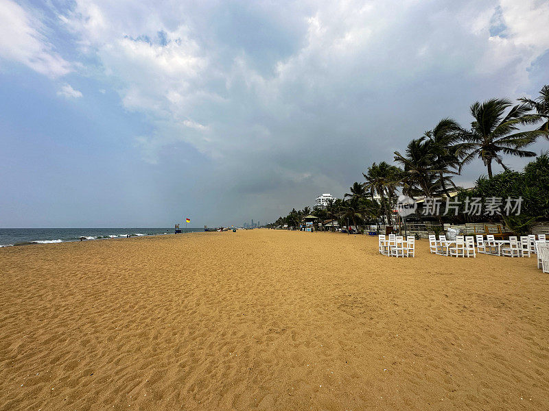 斯里兰卡，科伦坡，拉维尼亚山海滩的金色沙滩上的沙滩房屋、酒店和露天餐厅的图像，阳光穿过多云晴朗的蓝天，聚焦于前景
