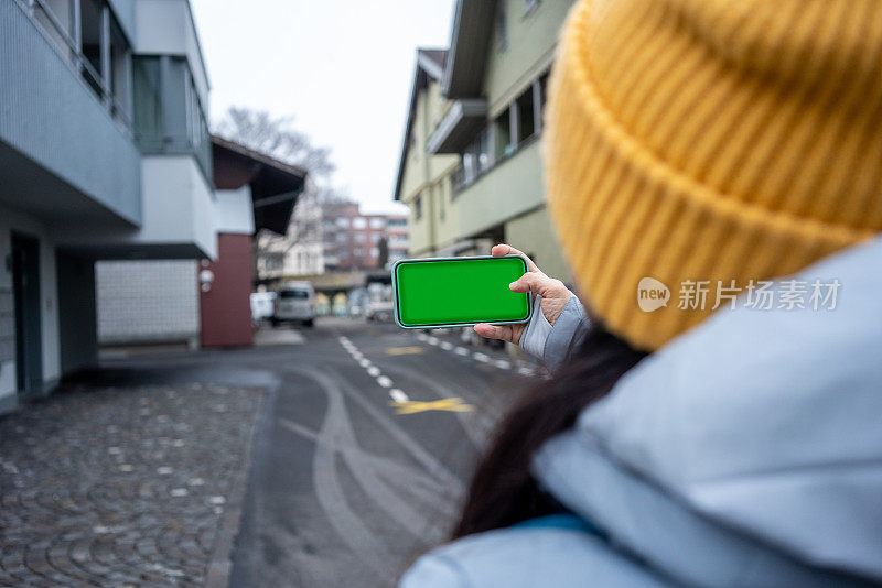 女人手里拿着绿屏的智能手机，她还在街头横行拍照