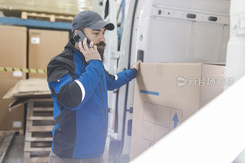 蓄着胡子的仓库工人在把包裹放进送货车的后备箱时使用了手机