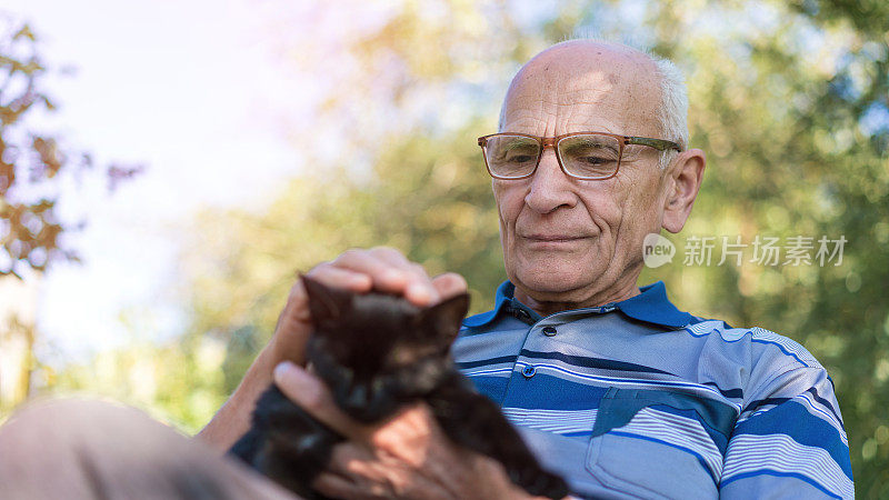 心满意足的老人戴着眼镜抱着小黑猫，在阳光明媚的花园里亲密无间。在宁静的环境中，温柔的陪伴和户外的放松