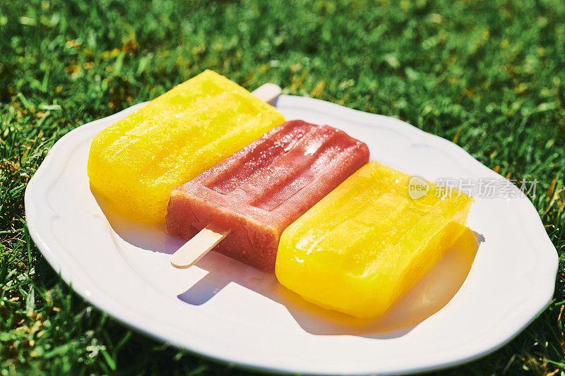 三根冰棍放在一个白色的盘子里，在一个炎热的阳光灿烂的夏天的花园草坪上。