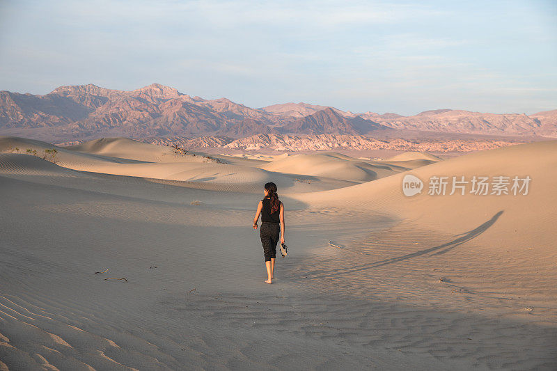 娇小的亚洲女人走过死亡谷的沙丘拿着凉鞋-强大的独立冒险旅行背景