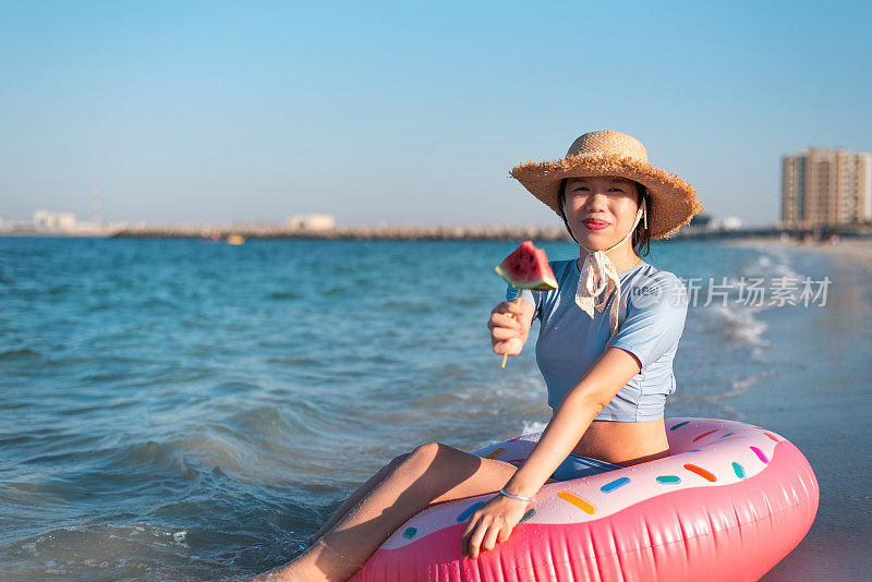 一个女人坐在沙滩上吃着西瓜，同时坐在一个漂浮的甜甜圈上