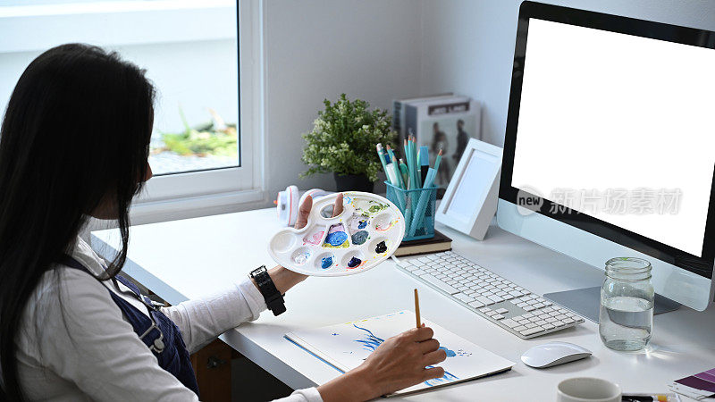 年轻的女艺术家在画室里用水彩画在纸上，而坐在她的电脑前。
