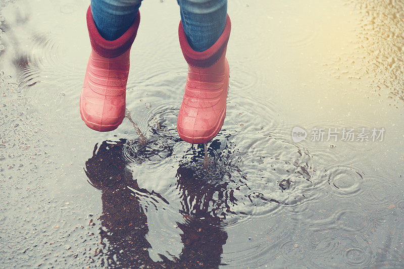 雨后穿着粉红色靴子的女孩在户外的水坑里跳。健美的。秋天的概念。