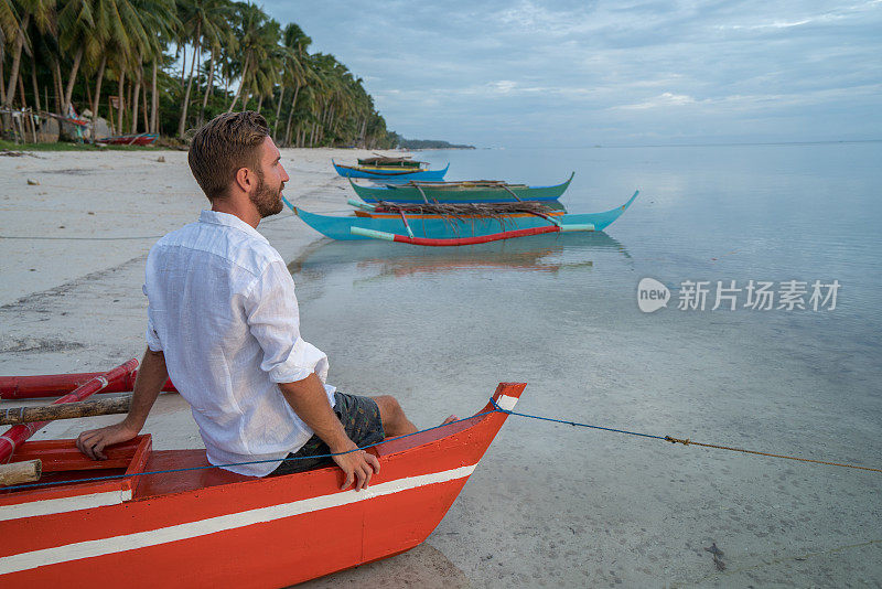 日落时分，一个年轻人坐在热带海滩上的船上，凝视着大自然