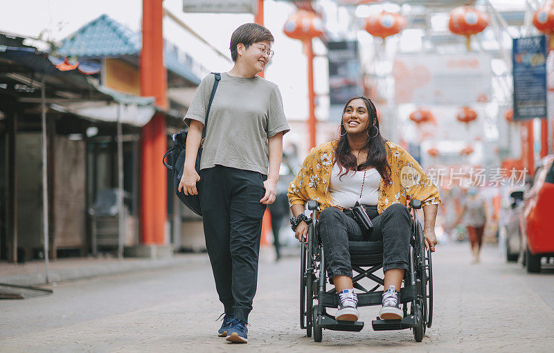 亚洲华人中年妇女和她的印度残疾女性朋友肩并肩走在城市街道上的轮椅上