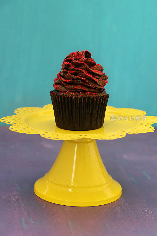 个人自制的棕色巧克力杯形蛋糕，纸制蛋糕盒放在黄色的蛋糕架上，巧克力黄油糖霜被脱水覆盆子粉末包裹，分割成灰色和蓝色背景，聚焦前景