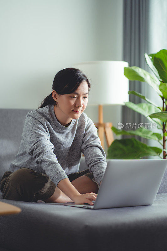 亚洲女性坐在家里的客厅里用笔记本电脑工作或上网。