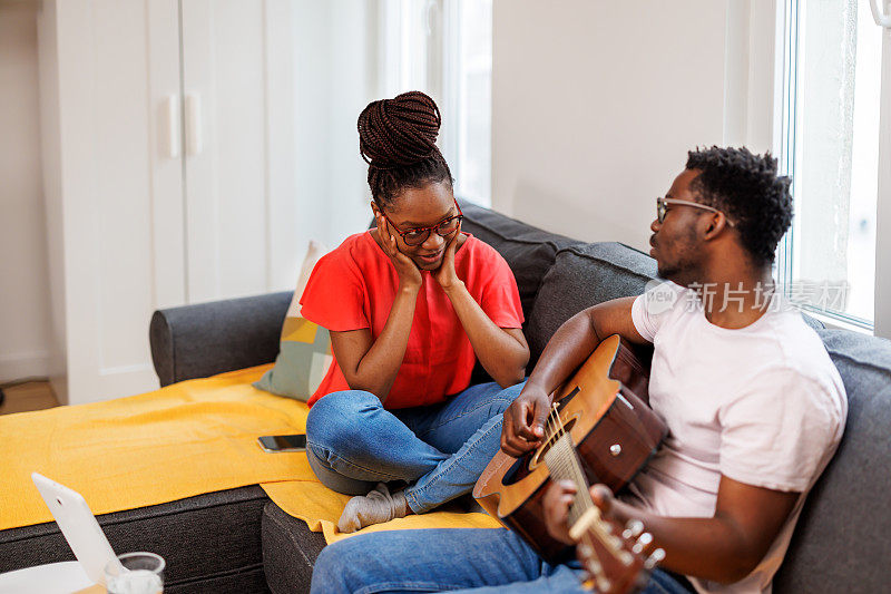 一位年轻的非洲男子在家中弹着木吉他，与女友一起享受着快乐的时光。