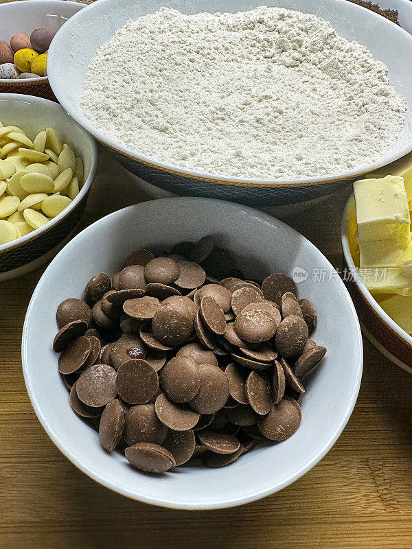 一组装有烘焙原料的白色碗的特写图像，大碗的白面粉，小碗的黄油块，糖果包裹的巧克力迷你复活节彩蛋，牛奶和白巧克力片，重点放在前景