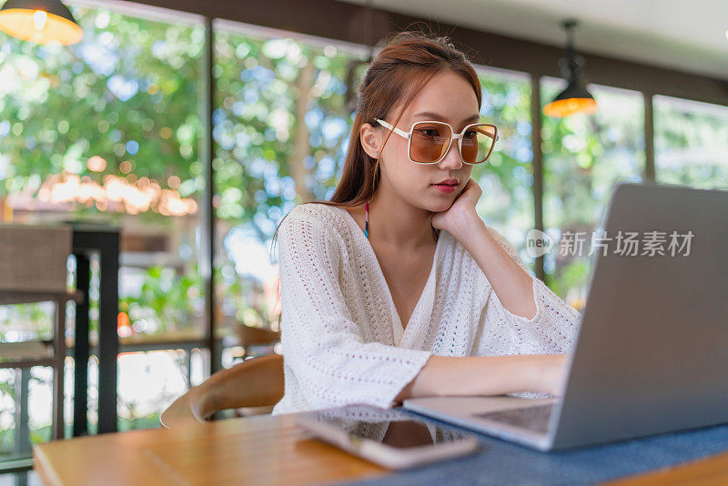 一名年轻的亚洲女子在海滩咖啡馆用笔记本电脑工作，身边放着一部智能手机。戴着太阳镜、穿着沙滩装的女性数字游民在度假氛围中摆弄着笔记本电脑。穿着沙滩装的亚洲女人坐在咖啡馆的沙发上用笔记本电脑远程工作。