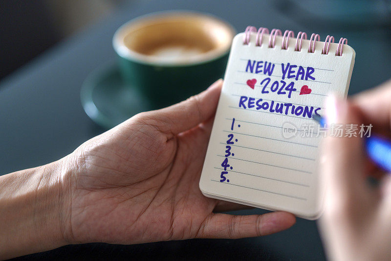 照片中，一位女性的手在工作台上的纸质笔记本上写下了新年决心和2024年的目标。