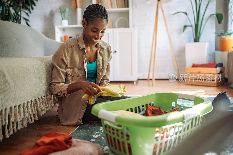 一位黑人妇女坐在舒适的客厅地板上，把要洗的衣服放在洗衣篮里分类。