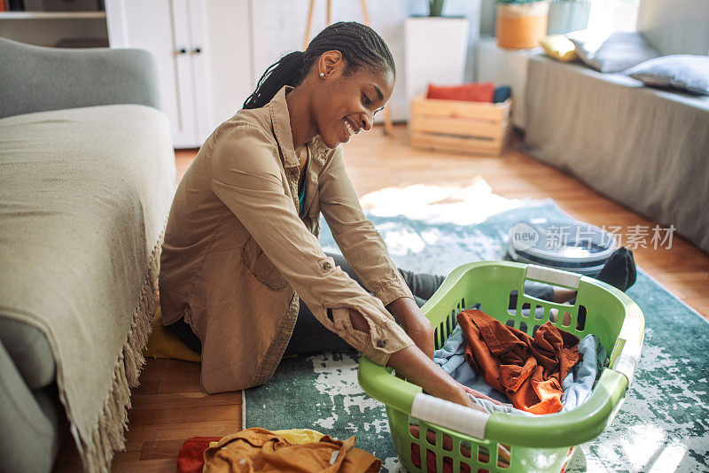 一位黑人妇女坐在舒适的客厅地板上，把要洗的衣服放在洗衣篮里分类。
