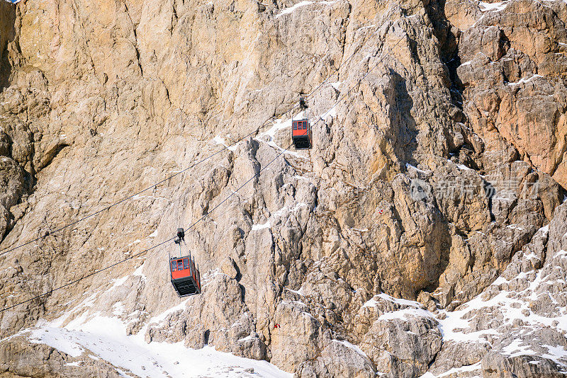 在一个阳光明媚的冬日，架空缆车客舱带着人们在白雪皑皑的岩石山顶上下穿梭