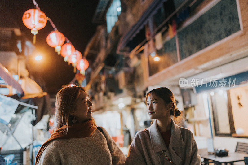 在香港，两名年轻的亚洲妇女在晚上肩并肩地走在一条似乎有商店的街道上，庆祝中国新年。
