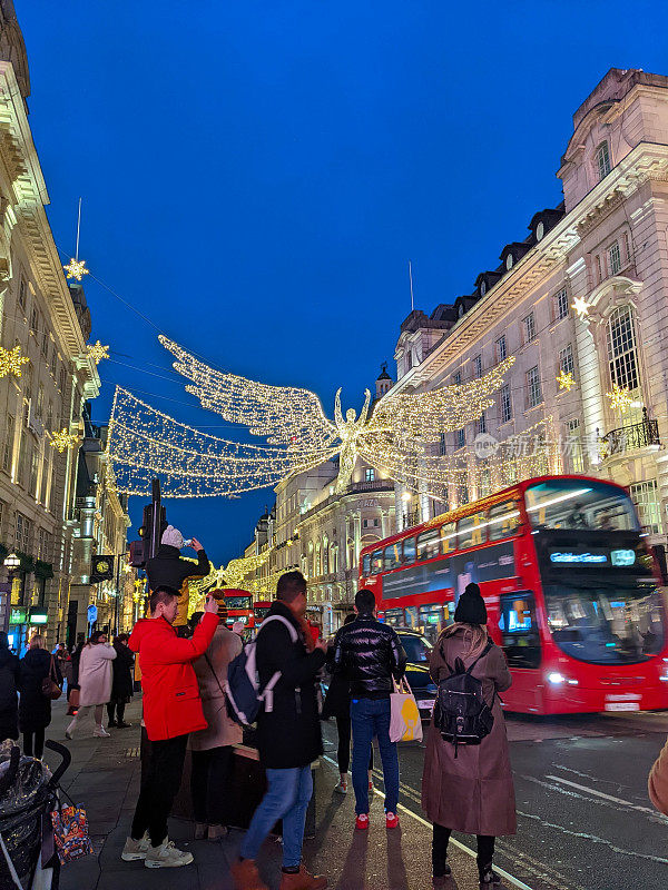 黄昏时分，行人在摄政街拍摄悬挂的天使圣诞彩灯装饰，汽车和一辆双层巴士经过