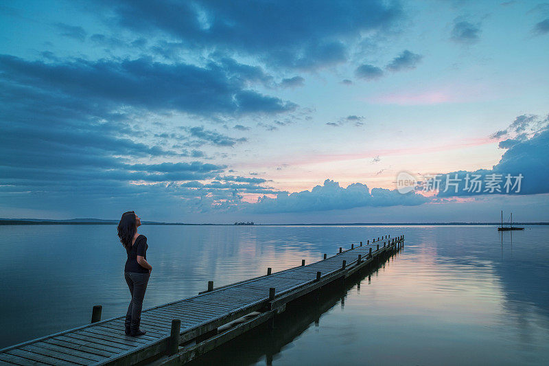 年轻女子在湖边的码头上观看雄伟的日落云景