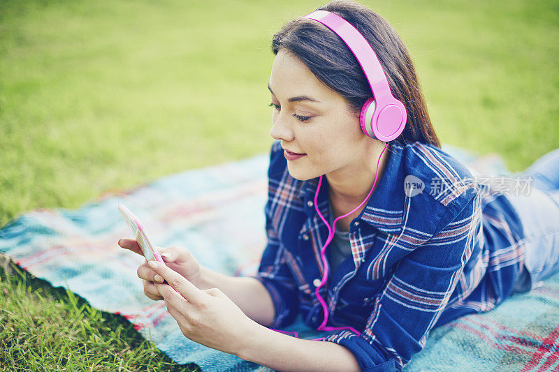 一个女人戴着耳机和智能手机躺在公园的毯子上