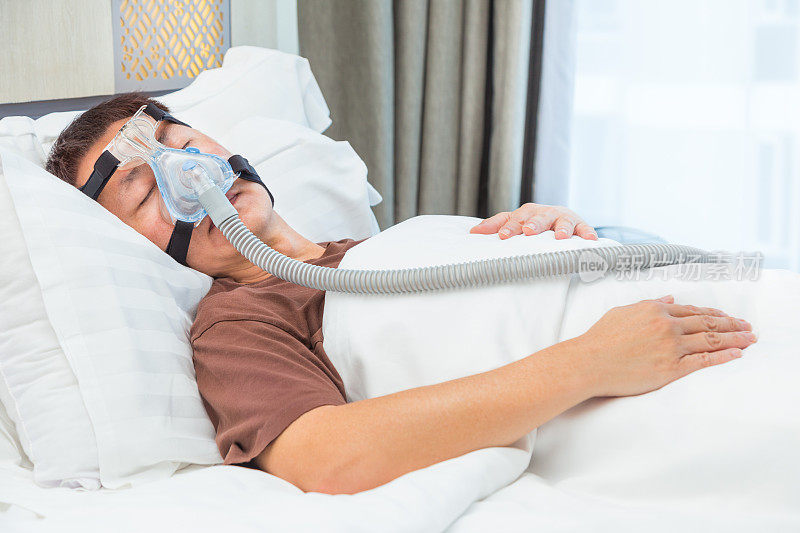 中年亚洲男子在床上睡觉，戴着连接空气软管的CPAP头罩，这是为患有睡眠呼吸暂停的人准备的设备