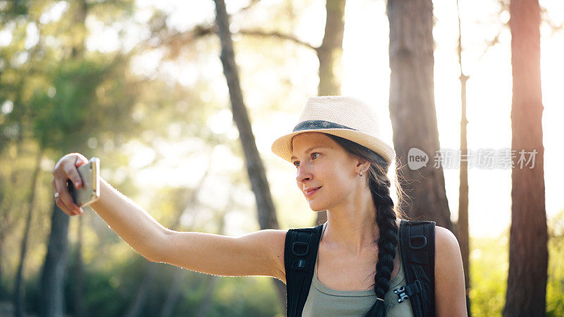 年轻的旅游女孩背包客在帽子在线视频聊天与智能手机在夏季森林散步