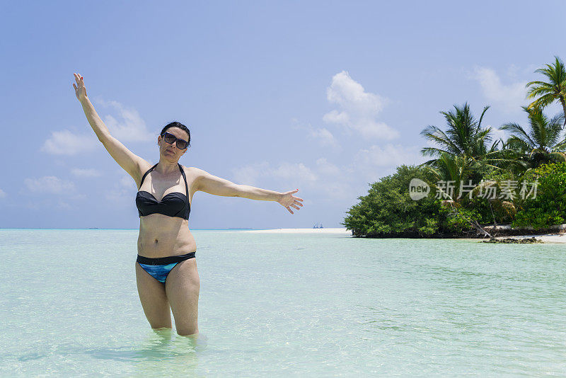 正面视图美丽的女人站在浅水与张开的手在小岛屿海滩在四月，马尔代夫