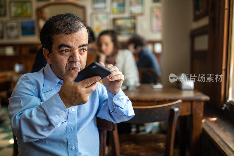 一个矮化的商人坐在当地的咖啡馆里用手机录制语音信息