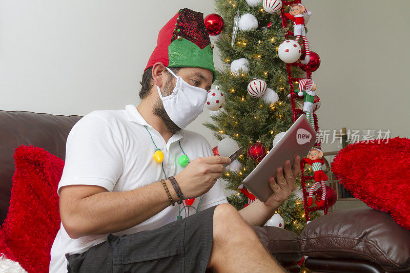 在冠状病毒Covid-19大流行疾病期间，一名年轻拉丁男子在装饰有圣诞树和装饰品的房子里崩溃购物和使用数字平板电脑和信用卡