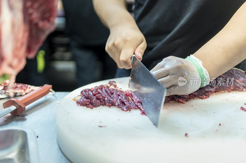 在一家牛肉火锅餐厅的开放式厨房里，厨师在挂着的切碎的生肉后面用刀切牛肉