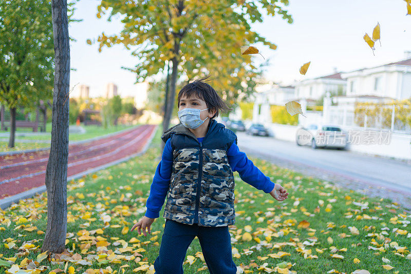 一个戴着防护面具的快乐小男孩正在公园里享受秋日落叶的乐趣。