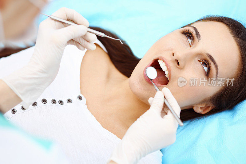 年轻的女病人正在看牙医。美丽的女人与健康的直白牙齿坐在牙科椅子与张大的嘴在口腔检查期间，而医生工作的牙齿