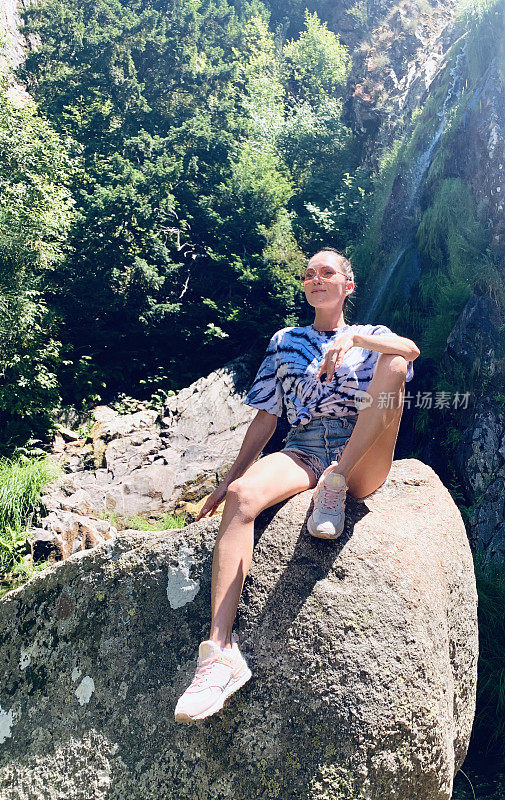 一个女孩坐在瀑布旁边的一块大石头上