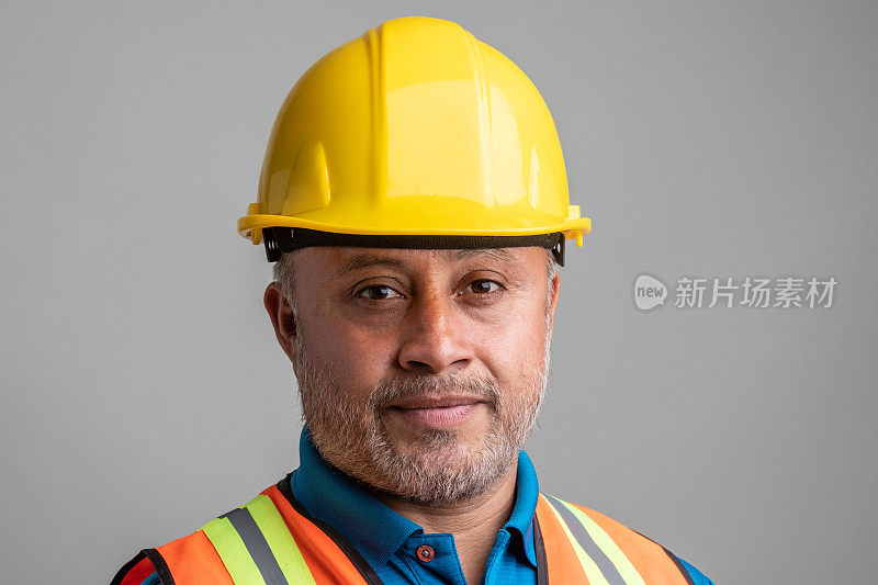 西班牙裔成年男性建筑工人戴着工作头盔，看着灰色背景上的摄像机