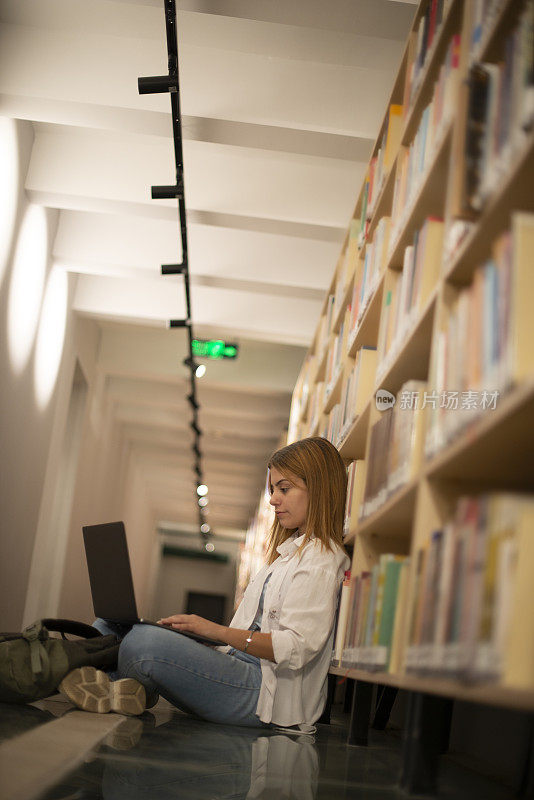 一名女学生倚靠在市图书馆的书架上，用笔记本电脑做研究