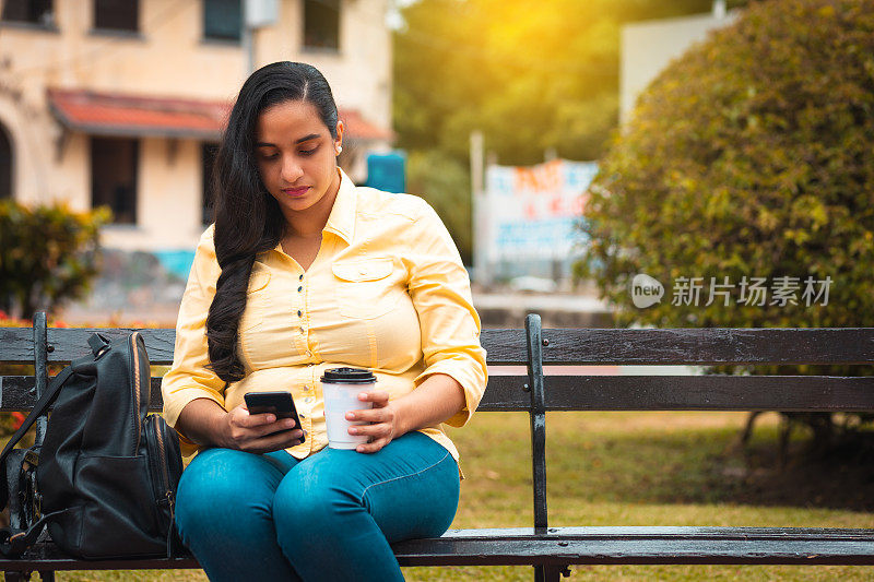 年轻的拉丁女人在公园喝咖啡时查看手机