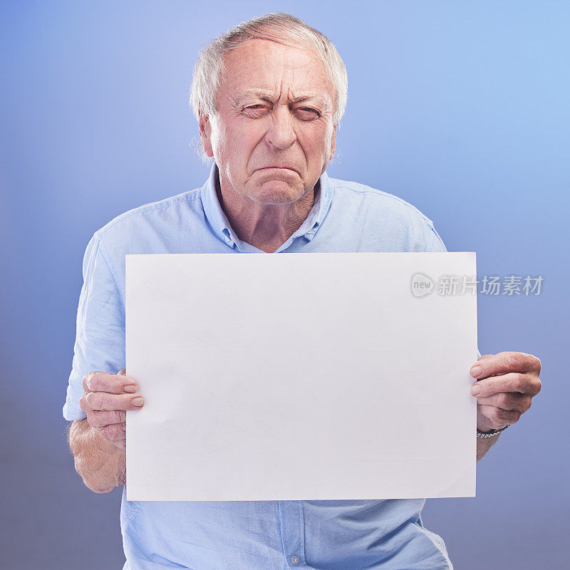 摄影棚拍摄的是一位年长的男子拿着一块空白的牌子，在蓝色的背景下看起来很不开心