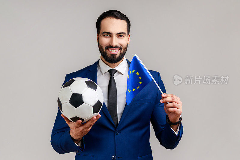 一名快乐的男子手持黑白相间的足球球和欧盟旗帜观看球迷最喜欢的球队。