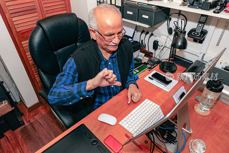 Bogotá，哥伦比亚-一个白发苍苍，74岁的亚洲印度人在他的家庭办公室在他的桌面电脑上工作。他做了一个不满意的表情，大拇指朝下的手势，表明他的工作进展不顺利。