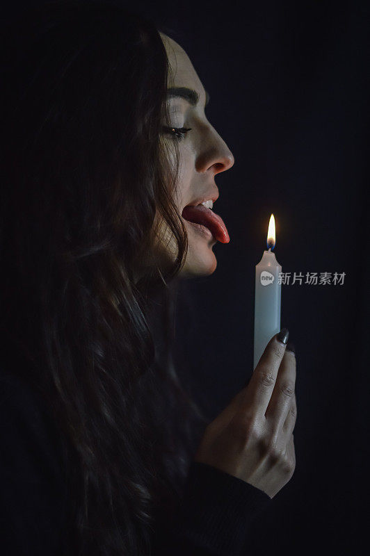 玩火——长发侧身的年轻美丽女子，手里拿着一根燃烧的蜡烛，假装舔着它的火焰